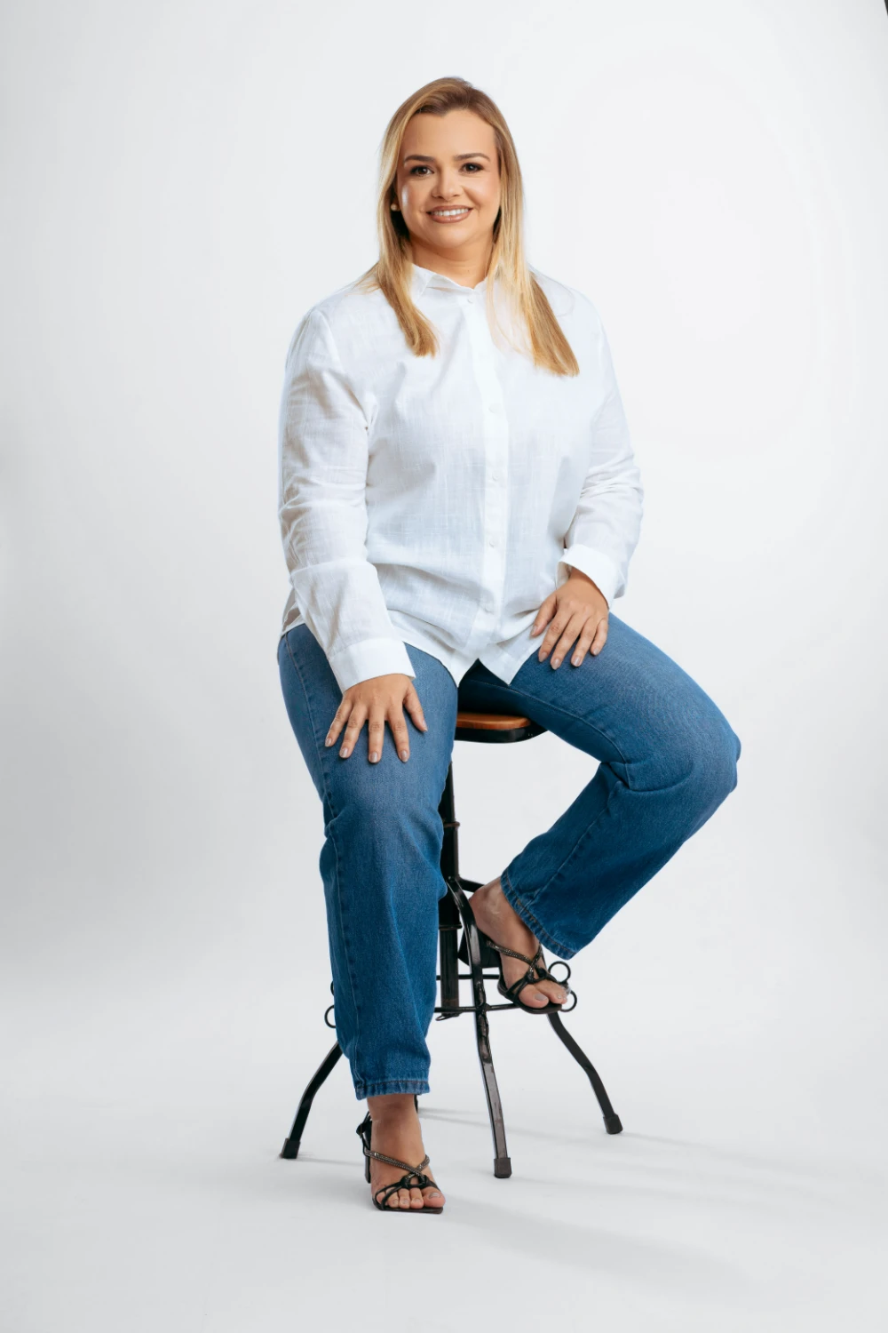 Paulinha Braga sentada em um banquinho, de camisa branca e calça jeans, sorrindo